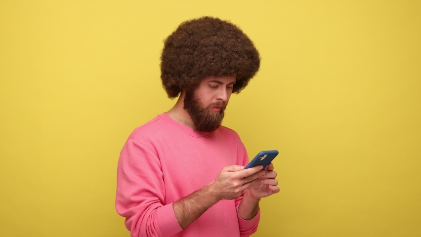 Hipster man with Afro hairstyle typing message on cell phone, chatting thinking over information, making choice, having doubts, wears pink sweatshirt. Indoor studio shot isolated on yellow background. Royalty-Free Stock Footage #1089057125
