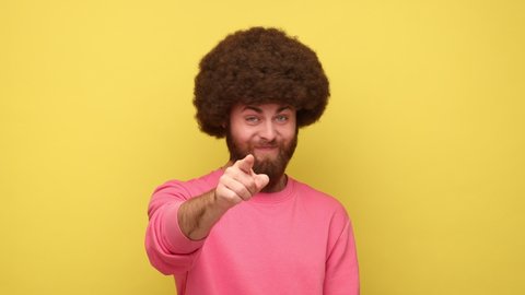 Bearded man looking with incredulous suspicious gaze and touching nose, gesturing you are liar, suspecting falsehood, wearing pink sweatshirt. Indoor studio shot isolated on yellow background.