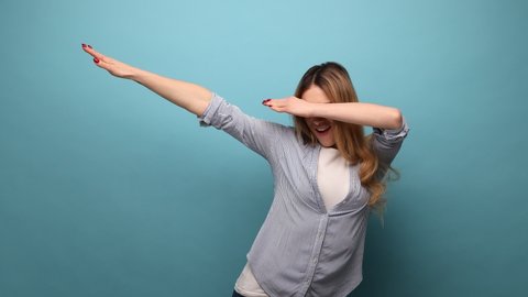 Portrait of attractive woman showing dab dance pose, famous internet meme of triumph, performing dabbing trends with hand gesture, wearing striped shirt. Indoor studio shot isolated on blue background