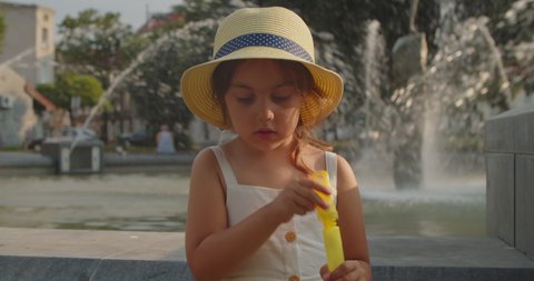 Cute preschool girl in a hat and dress blows soap bubbles against the background of a fountain in the city. Happy little girl is playing with soap bubbles in the summer on a sunny summer evening.
