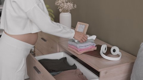 Cropped slowmo shot of expectant mother putting tiny baby clothes in drawer at home, stroking her belly with tenderness