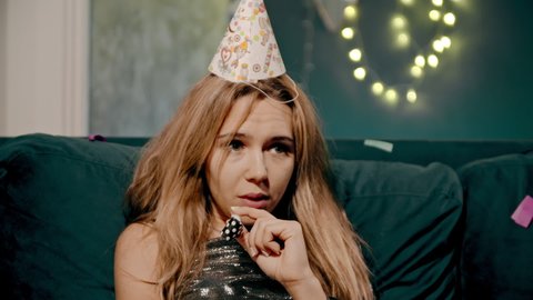 Close-up portrait of upset and sad lonely blonde girl lying or sitting on sofa or couch in birthday or festive cap and whistling whistle. Young woman in decorated house celebrating Party Party alone.