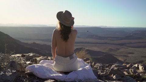 Young woman sitting topless on blanket at edge of Fish River Canyon in Namibia. Grand Canyon in USA. Rocky mountains, hiking. Travel in wild nature desert. Sunny summer day. Panoramic view viewpoint.