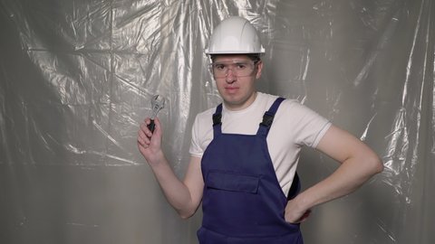 Construction worker. Builder in blue overalls and white hard hat, glasses with wrench. Building, repair and renovation of house or apartment. Man in helmet hold spanner on background plastic film.