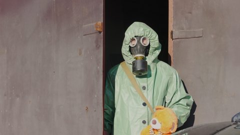 Soldier in anti nuclear costume and gas mask saving toy bear standing near metal door of shelter
