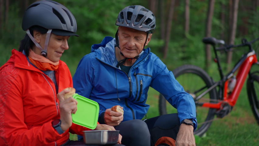 Happy senior couple bikers with eating snack outdoors in forest in autumn day.