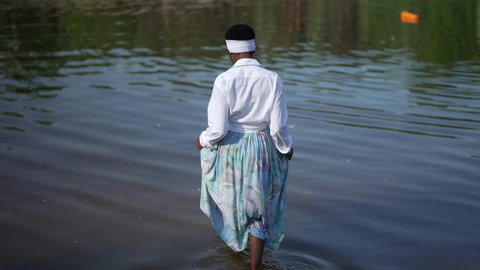 Back view slim African American young woman entering river stretching hands in slow motion. Wide shot relaxed barefoot lady in dress enjoying sunny day outdoors. Lifestyle and leisure