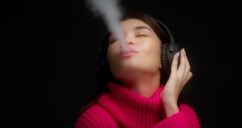 Close-up. Beautiful young woman in a pink sweater listens to music with wireless headphones, dances and smokes an electronic cigarette. Vaping smoke on a black background