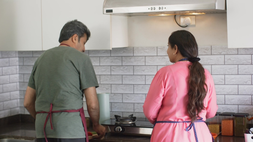 cheerful middle aged Indian Asian married couple or Husband and Wife wearing Chef Apron dancing, cherishing happy moments together in a modern kitchen house indoor. Healthy relationship concept  Royalty-Free Stock Footage #1089081419