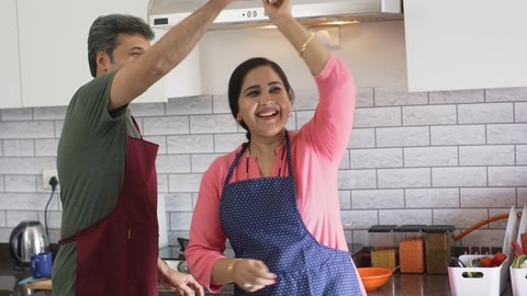 cheerful middle aged Indian Asian married couple or Husband and Wife wearing Chef Apron dancing, cherishing happy moments together in a modern kitchen house indoor. Healthy relationship concept : stockvideo