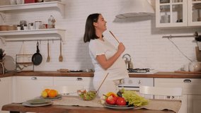 A cheerful pregnant woman has fun in the kitchen, she dances and sings holding a wooden spoon in her hands.
