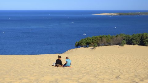 Dune of Pilat, La Teste-de-Buch, France - August 2021 : Multiracial couple sitting on the sand of the Dune of Pilat on the Arcachon Bay a summer day in Gironde, France