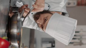 Vertical video: Male chef pouring shredded cheese on cooked food in frying pan, using grater and kitchen utensils to cook gourmet dish. Professional cook in white uniform adding grated parmesan on