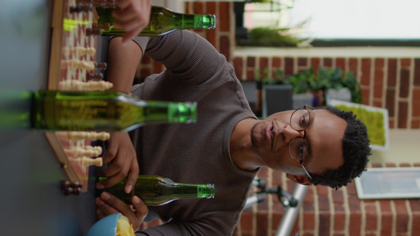 Vertical video: Portrait of african american man having fun with friends in living room, playing chess and board games. Young adult enjoying group gathering to play with beer and snacks for