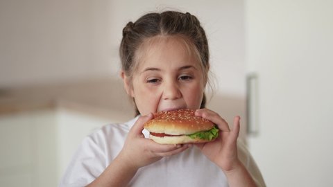 little girl eating a hamburger. unhealthy fast food proper nutrition concept. child greedily with pleasure bites a big burger in lifestyle the kitchen at home. kid eats fast food close-up