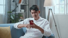 Smiling Asian Man Having Video Call On Smartphone While Lying On Sofa In The Living Room 
