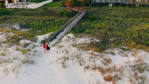 Two girls walking away from sandy beach on wooden walkway in dusk. Aerial view of mother and daughter walking landing foot bridge, returning home after having rest on beach. Concept of relaxation