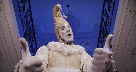A make-up cool dramatic funny mime guy on a blue theatrical stage shows thumbs up gesture. A theater actor dressed as an old Baroque harlequin. Copy space theme for promo video. Wow reaction