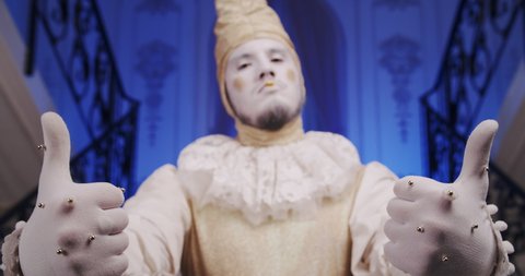A make-up cool dramatic funny mime guy on a blue theatrical stage shows thumbs up gesture. A theater actor dressed as an old Baroque harlequin. Copy space theme for promo video
