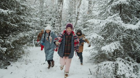 Joyful young friends smiling and running through snowy forest while having fun outdoors on winter day