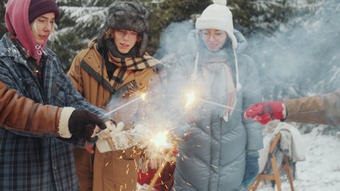 Company of young cheerful multiethnic friends lighting Christmas sparklers, smiling and jumping while celebrating holiday at campsite in winter forest