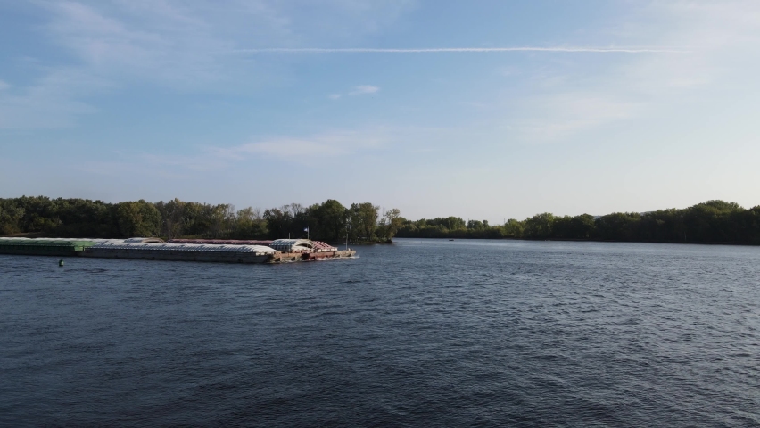 Empty barges being pushed up the Mississippi River on a bright summer day. Sun reflecting off the blue water. Groups of trees growing on islands in the river. Light clouds in the blue sky. 