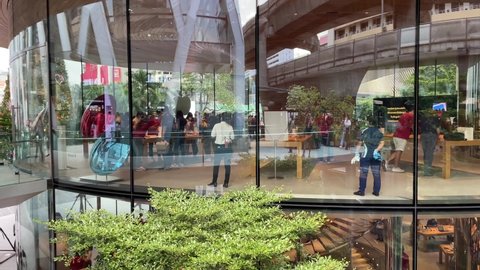 BANGKOK, THAILAND - Circa November, 2021: Apple Store at Central World, huge glass window view shopper inside. People walk in the entrance door. Sky train track in glass window reflection