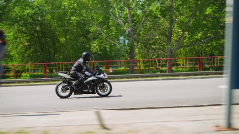Biker in black uniform and hardhat rides powerful motorcycle along large road past green park in city on sunny day side view slow motion