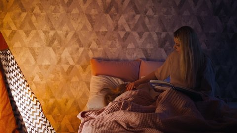 Long haired woman with book strokes head of little sleeping child covered with soft duvet near toy playhouse with glowing lamp in bedroom slow motion