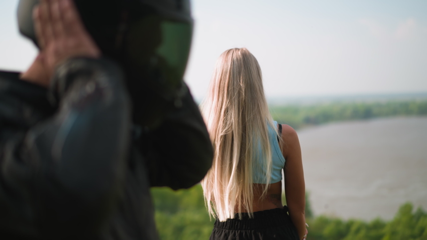 Long blonde haired woman enjoys view of large picturesque river while man biker takes off hardhat on hilly bank on spring day slow motion