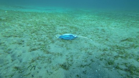 Used face mask lies on sandy bottom covered with green seagrass in sunlight. Discarded face masks polluting seabed since Coronavirus COVID-19. Pollution of Oceans. Camera moves forward, 4K-50fps