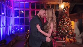 Man and woman in nice clothes having date in country house. Loving couple hugging and kissing at the backdrop of decorated table and Christmas tree.