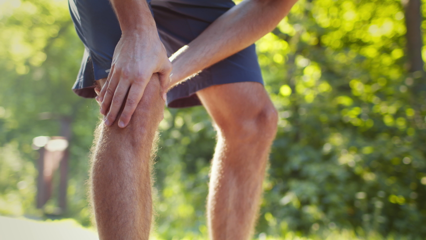 Sport and traumas concept. Unrecognizable man jogger running and feeling acute pain in knee, massaging painful leg during morning run in park, close up, slow motion, free space | Shutterstock HD Video #1089109521