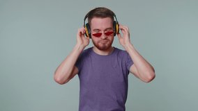 Portrait of bearded man 20s years old in purple t-shirt listening music via headphones and dancing disco fooling around having fun expressive gesticulating hands. Young one guy on gray wall background