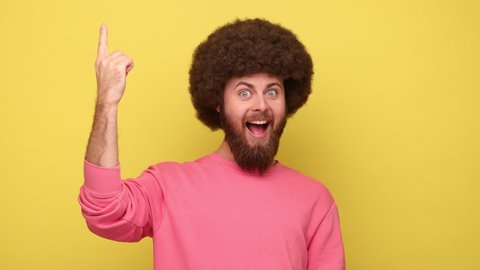 Bearded man with Afro hairstyle raising finger having genius idea, looking amazed by sudden smart solution, creative thought, wearing pink sweatshirt. Indoor studio shot isolated on yellow background.
