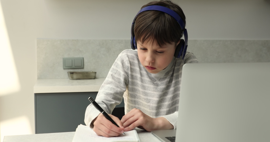 Distant learning. Focused preteen schoolboy in wireless headphones sit at home workplace by laptop take notes of video class lesson. Tween boy studying remotely engaged in elearning via conference app Royalty-Free Stock Footage #1089117005