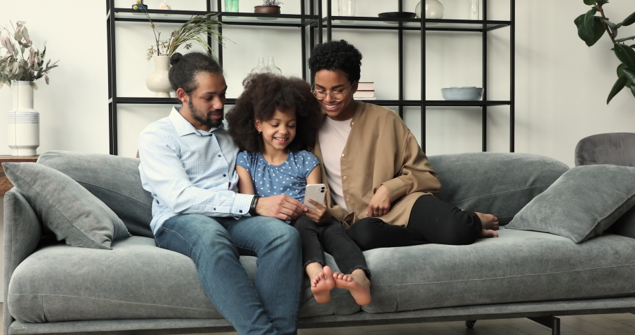 Loving African family of 3 mom dad little daughter schoolgirl relax on comfy couch hold cellphone browse websites. Friendly couple with tween daughter use smartphone do shopping order tickets for trip