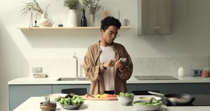 Pretty Black woman take pause in cooking dietary meal from fresh vegetables browse social media on cell. Young lady modern day housewife enjoy culinary at home kitchen use phone app to choose recipe