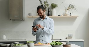 Smiling young African American man cooking food stand by kitchen table hold mobile phone ask professional advice in culinary chat. Millennial man chef search delicious recipe at web online using cell