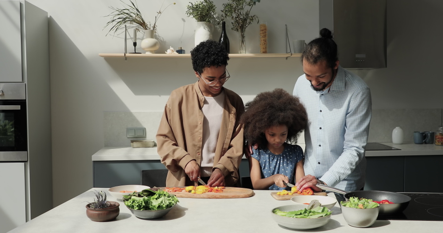 Friendly Black family young parents preteen child daughter prepare lunch dinner cut vegetables for healthy meal. Smiling dad mom teach little girl to make food at home praise hug kid learning to cook Royalty-Free Stock Footage #1089117249