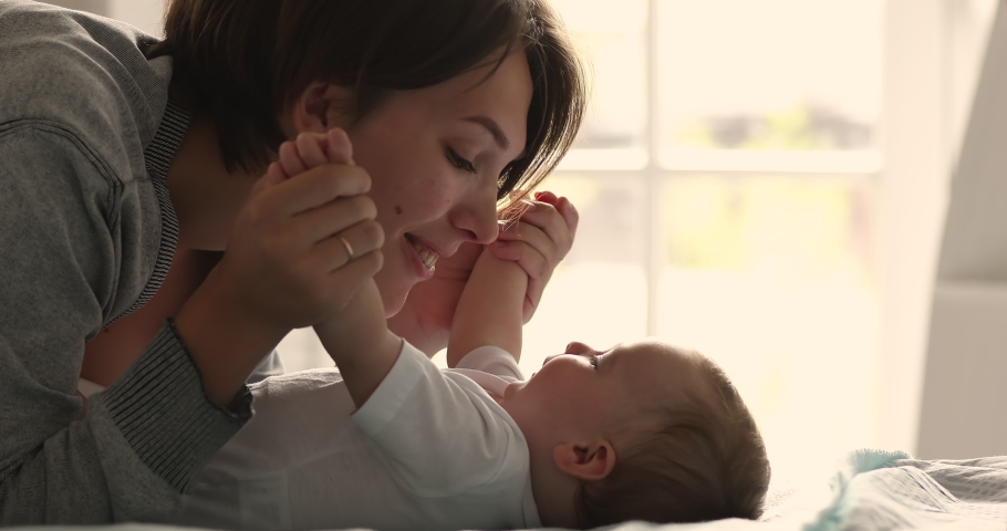 Caring young mommy enjoy playing with baby lying on back on bed touch kiss tiny nose plumpy cheeks whisper tender words. Carefree small infant laugh look in mom eyes receive caress from beloved parent Royalty-Free Stock Footage #1089117279