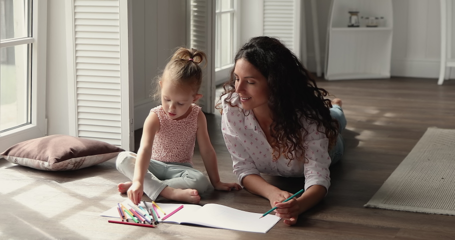Loving millennial Latin foster mom relax on warm floor with small kid adopted daughter engaged in drawing create fairy tale in pictures. Mother and child girl coloring sketches in album using pencils Royalty-Free Stock Footage #1089117283