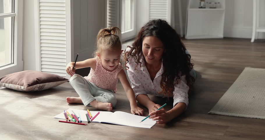 Loving millennial Latin foster mom relax on warm floor with small kid adopted daughter engaged in drawing create fairy tale in pictures. Mother and child girl coloring sketches in album using pencils | Shutterstock HD Video #1089117283