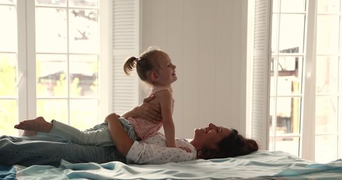 Happy little child girl preschooler cuddle with mom on soft large double bed play funny game smile talk. Serene Hispanic mother embrace hold in arms small daughter enjoy good morning in bright bedroom