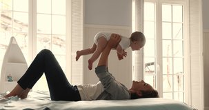 Happy loving millennial mom lie on back on soft big bed in light cozy bedroom lift up toss in air small baby daughter or son. Caring mum laugh cuddle kiss cute infant babe have fun enjoy motherhood