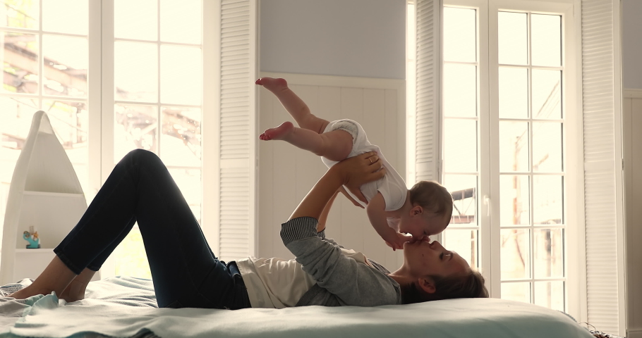 Happy loving millennial mom lie on back on soft big bed in light cozy bedroom lift up toss in air small baby daughter or son. Caring mum laugh cuddle kiss cute infant babe have fun enjoy motherhood | Shutterstock HD Video #1089117309