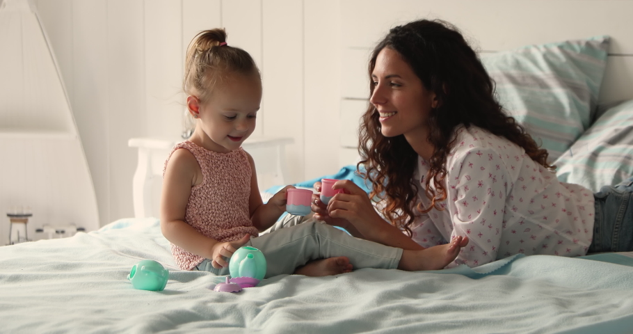 Friendly multiethnic family Latina mommy Caucasian daughter playing toy teaset on comfy bed with orthopedic mattress. Happy mom small child girl pretend tea party drink imaginary coffee from toy cups Royalty-Free Stock Footage #1089117323