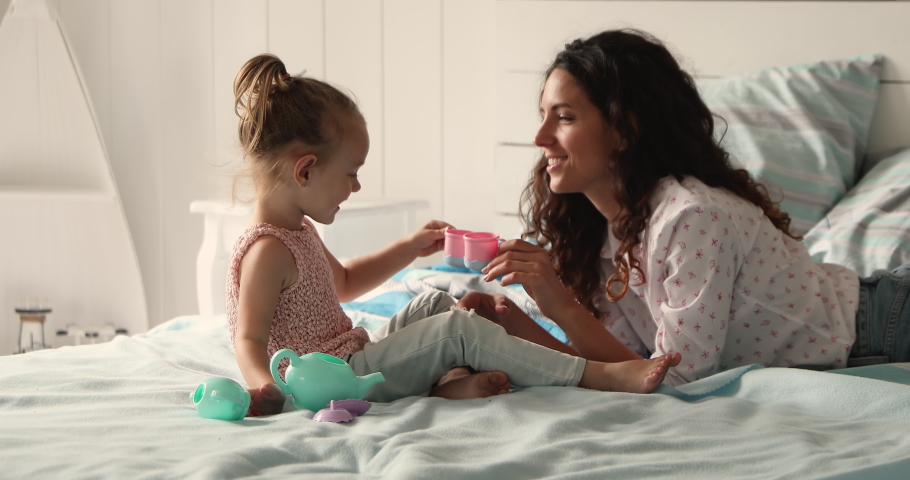 Friendly multiethnic family Latina mommy Caucasian daughter playing toy teaset on comfy bed with orthopedic mattress. Happy mom small child girl pretend tea party drink imaginary coffee from toy cups Royalty-Free Stock Footage #1089117323