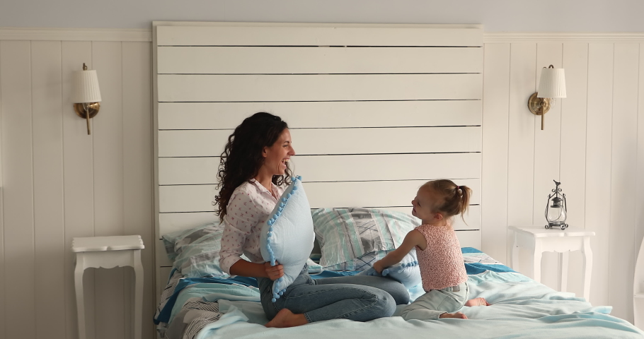 Energetic laughing Latin foster mom European adopted kid daughter enjoy funny pillow fight on big cozy bed mess have fun together. Joyful mother child girl play hilarious combat on soft firm mattress Royalty-Free Stock Footage #1089117327