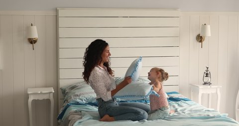 Energetic laughing Latin foster mom European adopted kid daughter enjoy funny pillow fight on big cozy bed mess have fun together. Joyful mother child girl play hilarious combat on soft firm mattress